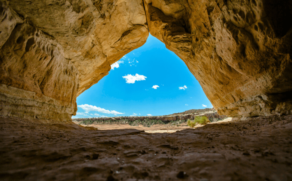 View from inside a rocky cave looking out at a bright blue sky with scattered clouds and desert landscape in the distance. The cave walls frame the opening in a roughly triangular shape—Request a Demo to See Radiant Logic In Action.