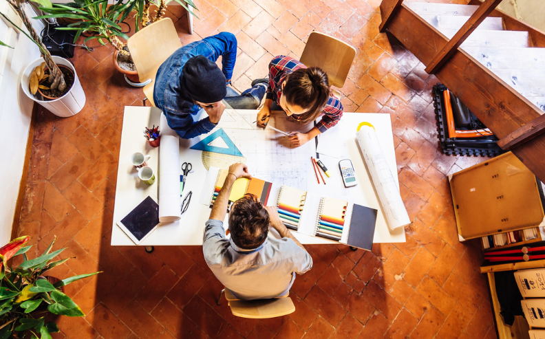 Three people sit around a table covered with pencils, blueprints, and stationery, collaborating on a project in a bright workspace with plants—focused on optimizing user access audit and compliance using automated tools.