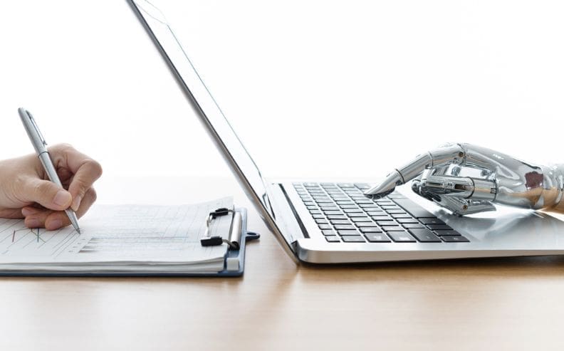 A human hand writes on a clipboard with a pen while a robotic hand types on a laptop keyboard beside it, symbolizing collaboration between humans and artificial intelligence.