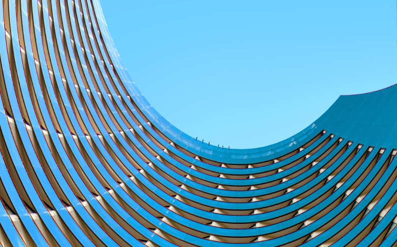 Curved, modern building with repeating horizontal lines and glass panels, forming a U-shape against a clear blue sky. The architecture creates a dynamic, abstract pattern.