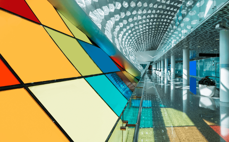 A modern building interior with colorful geometric glass panels on the left and a long corridor with white columns, patterned ceiling, and reflections on the shiny floor.