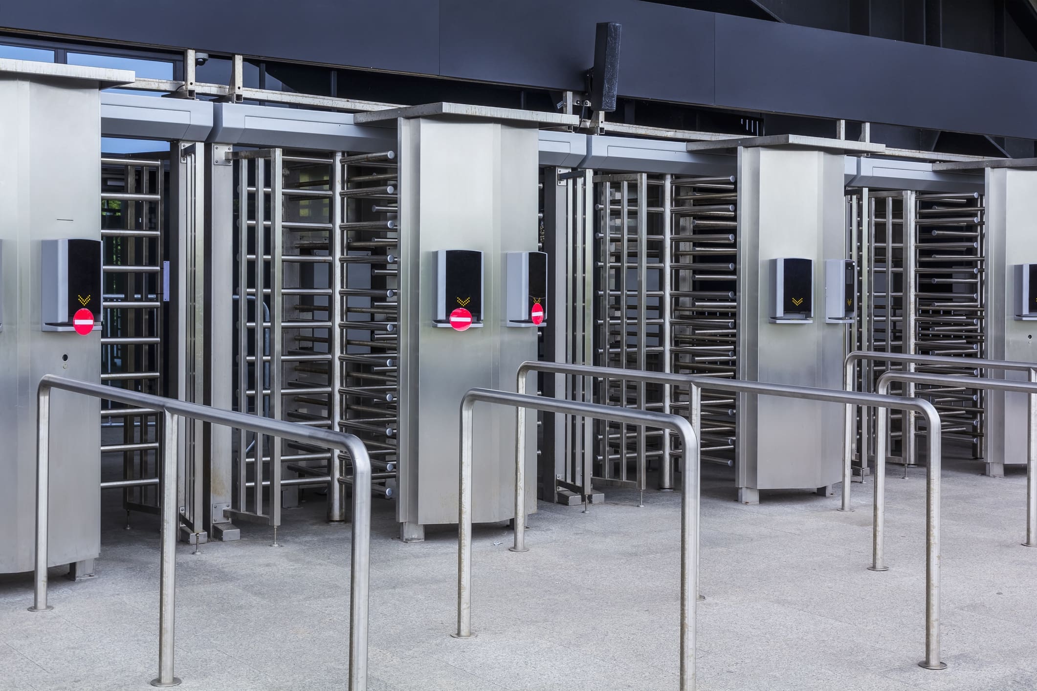 A row of modern, stainless steel full-height turnstiles with barriers, card readers, and red no-entry signs, set at an entrance or exit point, possibly for secure access to a building or venue.