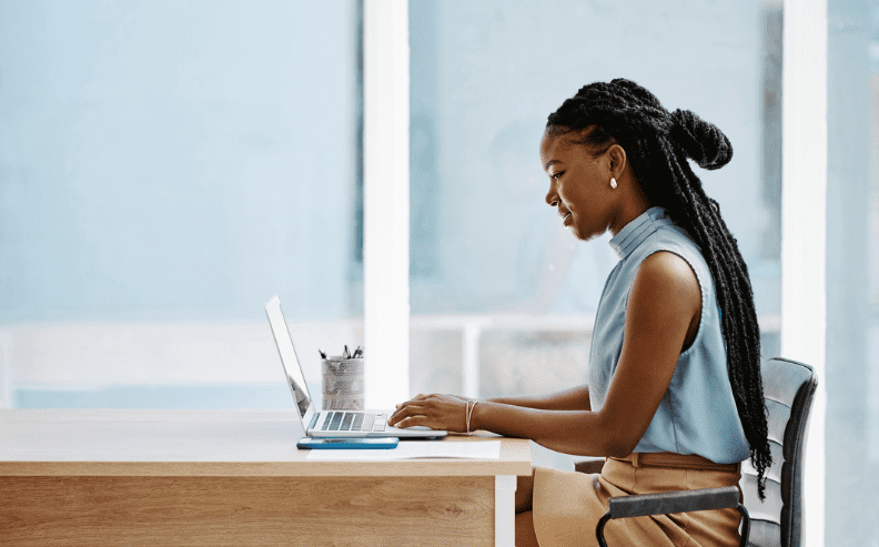 A woman with long braids sits at a desk, working on a laptop. She is wearing a sleeveless blue blouse and tan skirt, and the background is bright with large windows.
