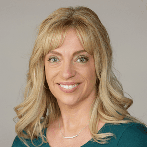 A woman with long, wavy blonde hair and light eyes smiles at the camera. She is wearing a teal top and a delicate necklace, with a neutral-colored background.
