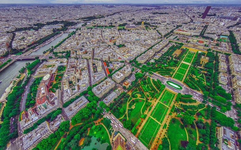 Aerial view of Paris, showing dense city buildings, the River Seine, and the green lawns of Champ de Mars park leading to the Eiffel Tower (not visible), with cloudy skies overhead.