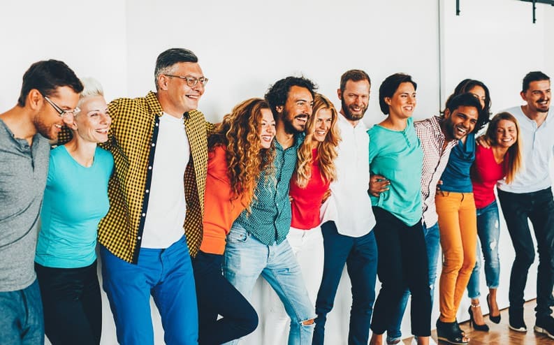 A diverse group of people stand in a row with arms around each other’s shoulders, smiling and laughing, in a bright indoor setting with a plain white wall background.