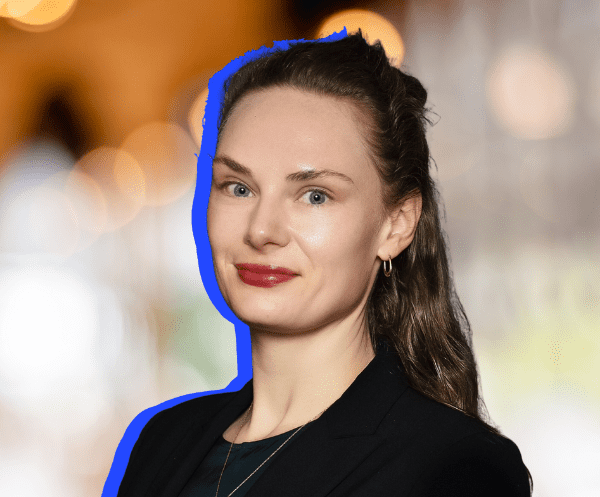 A woman with light skin and long brown hair, wearing a black blazer, smiles slightly at the camera. The background is blurred with warm lights, and a blue outline highlights her figure.