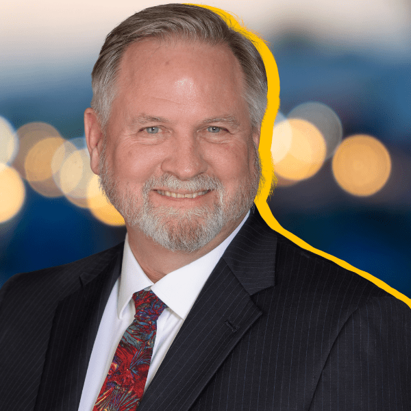 A smiling man with gray hair and beard, wearing a dark pinstripe suit, white shirt, and red patterned tie, stands against a blurred background with yellow bokeh lights and a yellow outline around him.