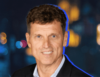 A man with short brown hair wearing a dark suit jacket and light blue shirt, smiling at the camera. The background is dark and blurred with colorful lights.