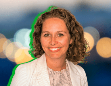 A smiling woman with curly brown hair, wearing a white blazer and a light patterned blouse, stands in front of a blurred background with colorful bokeh lights.