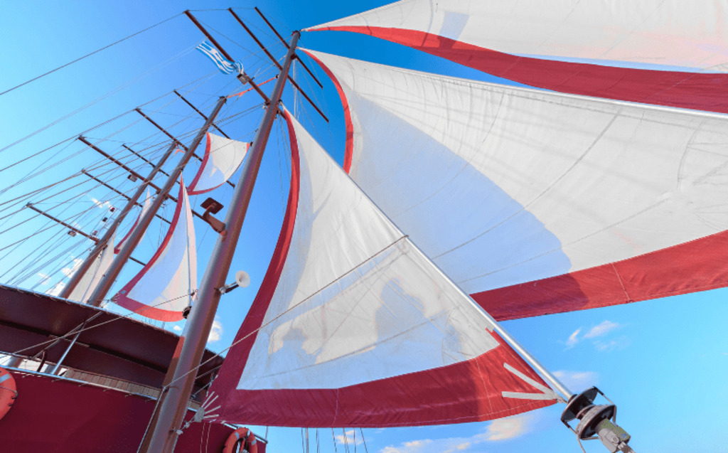 Bright white and red sails of a large sailboat or ship billow against a clear blue sky, viewed from below; part of the deck and rigging are visible, and a small flag is flying from a mast.