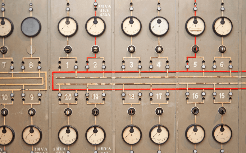 A close-up of an old-fashioned control panel with multiple analog gauges, switches, and labeled dials. Red and yellow circuit lines connect the numbered indicators across the panel.