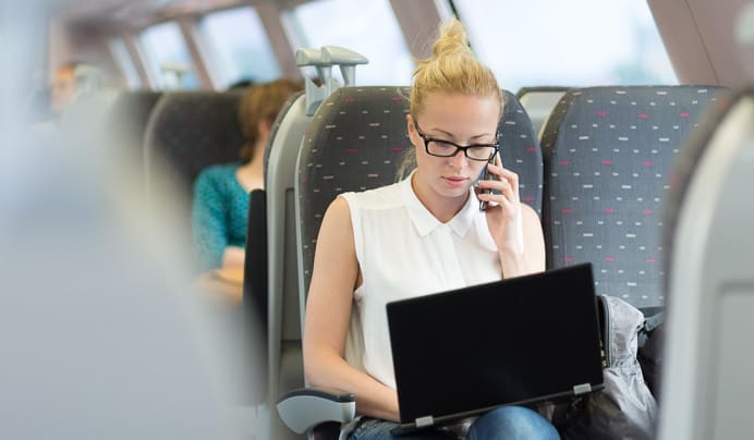 A woman with glasses and blonde hair tied up is sitting on a train, talking on her phone while working on a laptop. Other passengers are seated in the background.