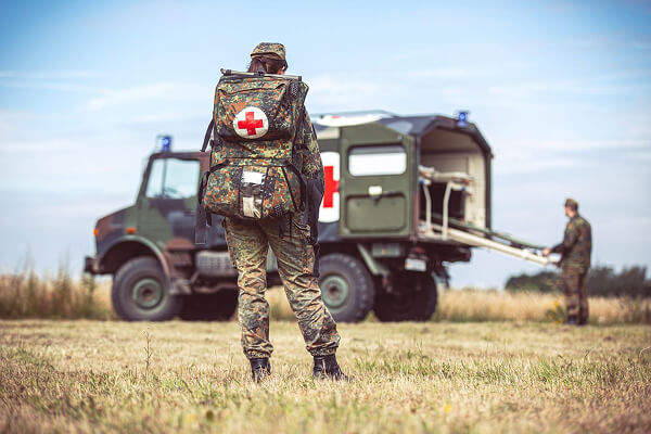 A person in military uniform with a medical backpack stands on grass facing an ambulance truck, while another person loads a stretcher into the vehicle in the background.