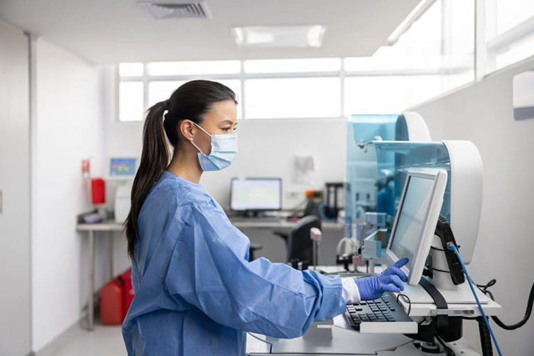 A woman wearing a mask, gloves, and a blue medical gown operates a touchscreen computer in a bright, modern laboratory with various scientific equipment in the background.