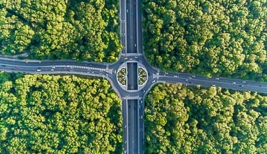 Aerial view of a roundabout with four roads intersecting in the middle of a dense green forest, with cars visible on the roads and the roundabout featuring a landscaped circular garden.