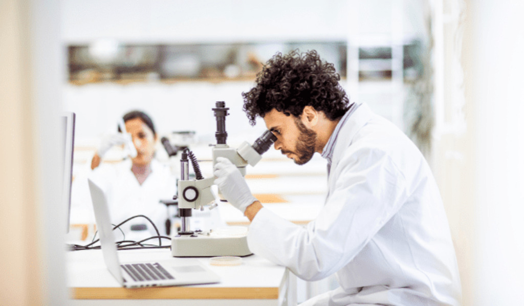 A scientist in a white lab coat and gloves looks into a microscope at a laboratory desk with a laptop. Another person in a lab coat works in the background.