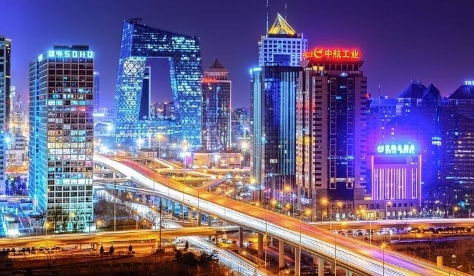 Night view of a modern city with brightly lit skyscrapers, colorful neon signs, and a busy highway with light trails from moving vehicles. The unique CCTV Headquarters building is visible among the high-rises.