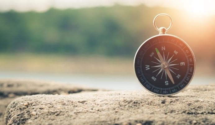 A black and silver compass resting on a rough stone surface outdoors, with blurred green trees and bright sunlight in the background.