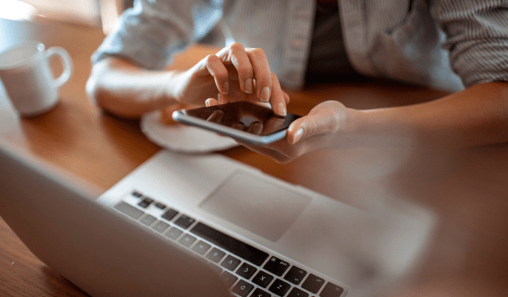 A person uses a smartphone while sitting at a desk with an open laptop and a coffee mug, suggesting multitasking or remote work in a casual setting.