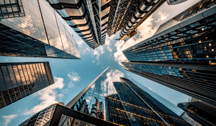 Looking up at modern skyscrapers with glass and steel facades reflecting the sky and clouds, creating a dynamic urban scene framed by tall buildings.