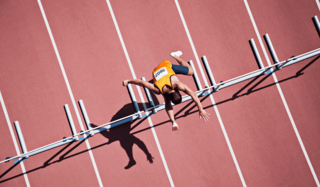 An athlete in yellow hurdles uniform trips and falls mid-race, arms outstretched, on a red running track with white lines and hurdles.