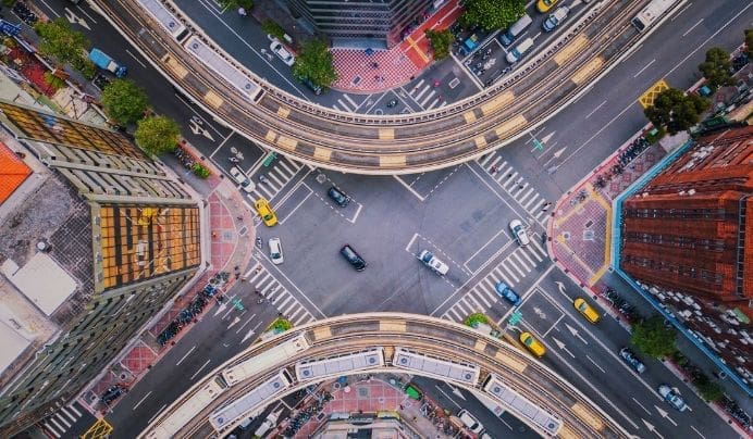 Aerial view of a busy urban intersection with elevated train tracks curving above the streets, cars moving in multiple directions, and tall buildings surrounding the area.