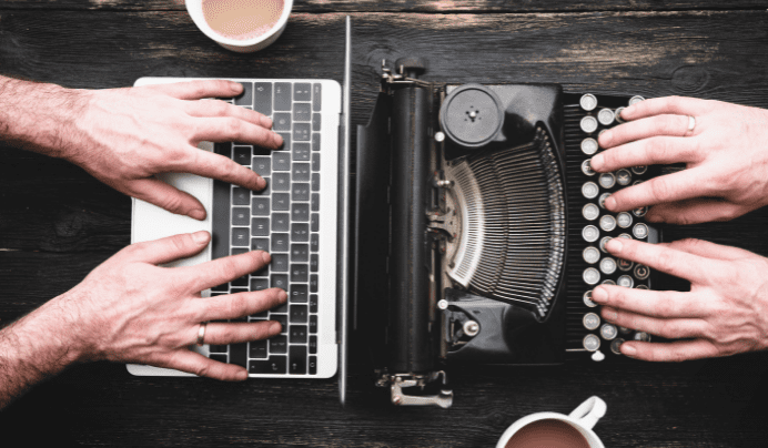 Two people typing side by side, one on a laptop keyboard and the other on a vintage typewriter, with two cups of coffee on a dark wooden table.