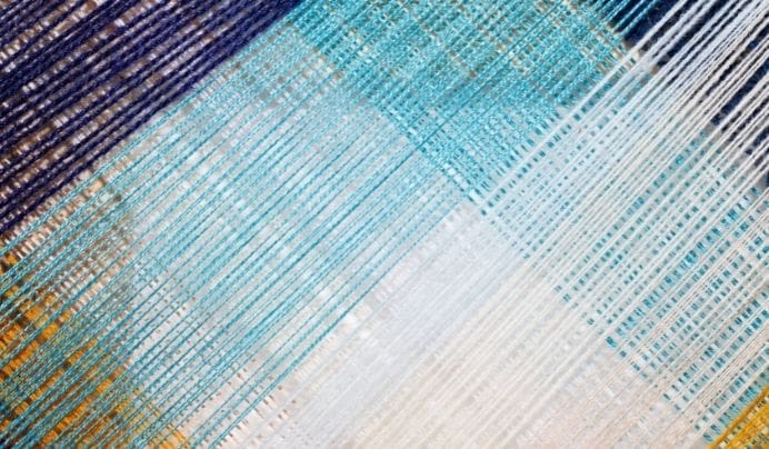 Close-up of multiple yarn threads in shades of blue, white, and yellow, arranged diagonally and overlapping in a geometric pattern. The threads create a textured and colorful woven appearance.
