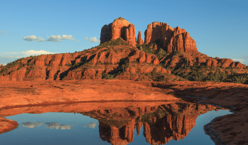 Red rock formations rise above a calm pool of water, reflecting the rocks and blue sky. Green vegetation dots the landscape under warm sunlight, creating a dramatic desert scene.