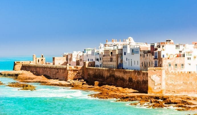 A coastal cityscape with white buildings and a fortified wall along the shoreline, bordered by turquoise-blue sea under a clear, bright sky.