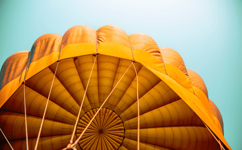 A close-up view from below of a bright yellow-orange parachute canopy against a clear blue sky, with suspension lines visible converging at the center.