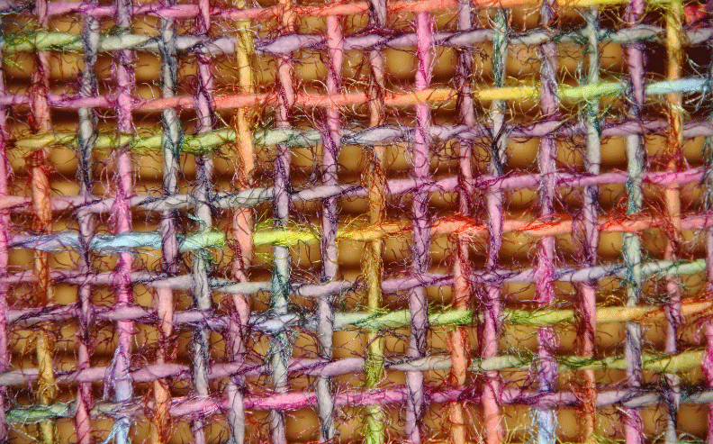 Close-up view of a colorful woven fabric, showing interlaced threads in pink, purple, yellow, green, orange, and blue, creating a textured, grid-like pattern with visible fibers and gaps between threads.