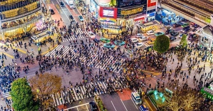 Aerial view of a busy city intersection with large crowds of people crossing in multiple directions, surrounded by tall buildings, bright lights, and many cars on the streets.