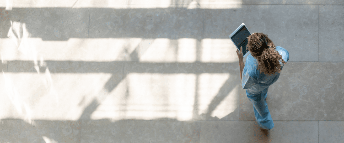 A person in blue scrubs walks across a sunlit floor while looking at a tablet, with long shadows cast on the light-colored tiles.