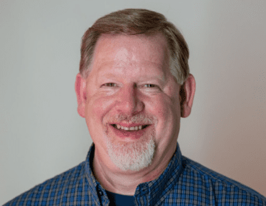 A middle-aged man with short light brown hair and a goatee smiles at the camera. He is wearing a blue plaid shirt and standing in front of a plain, light-colored background.