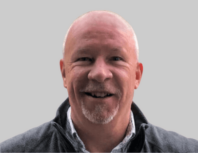 A middle-aged man with a bald head and short white beard, smiling. He is wearing a gray jacket over a light-colored collared shirt, and the background is plain gray.