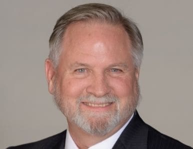A middle-aged man with gray hair and a beard smiles while wearing a dark suit and white shirt, set against a plain, light-colored background.