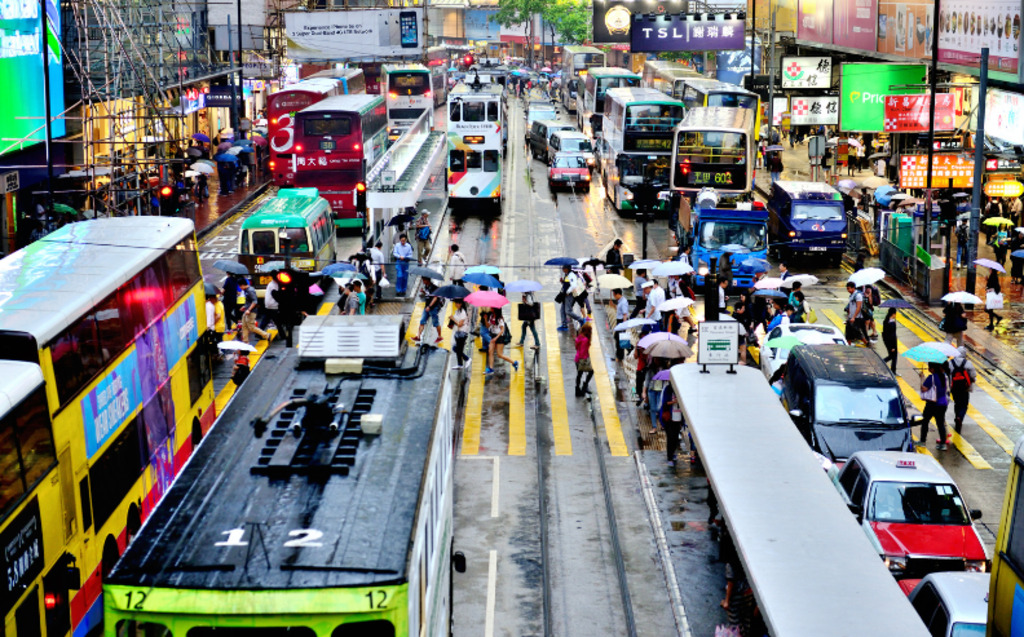 Busy city street with double-decker buses, cars, and people crossing in the rain holding colorful umbrellas. Bright lights and signs line the buildings. The scene is lively and bustling despite the wet weather.