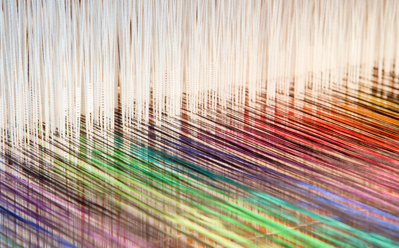 Close-up of colorful threads woven through the vertical strands of a loom, showing an array of vibrant colors including green, blue, red, and orange, creating an intricate textile pattern.