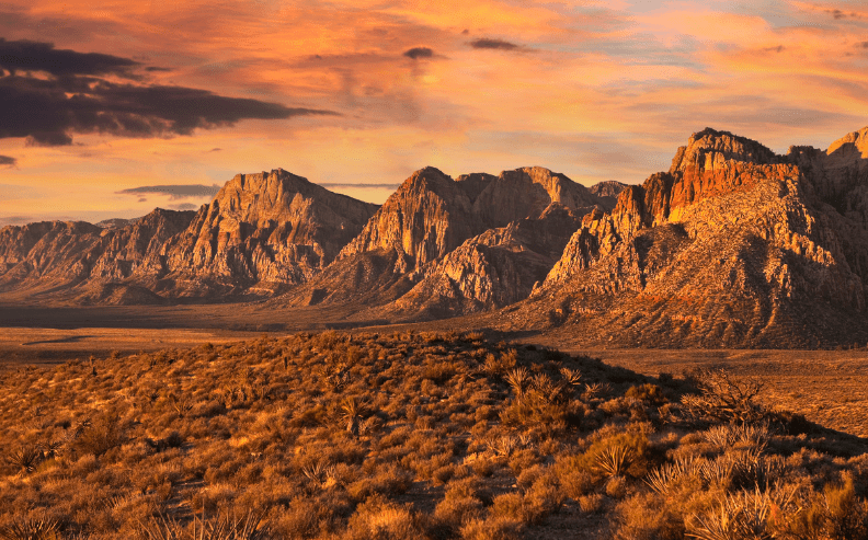 Desert landscape with rugged mountains under a dramatic orange sky at sunset. The foreground features sparse vegetation and dry, rocky terrain, while the distant peaks are illuminated by warm sunlight.