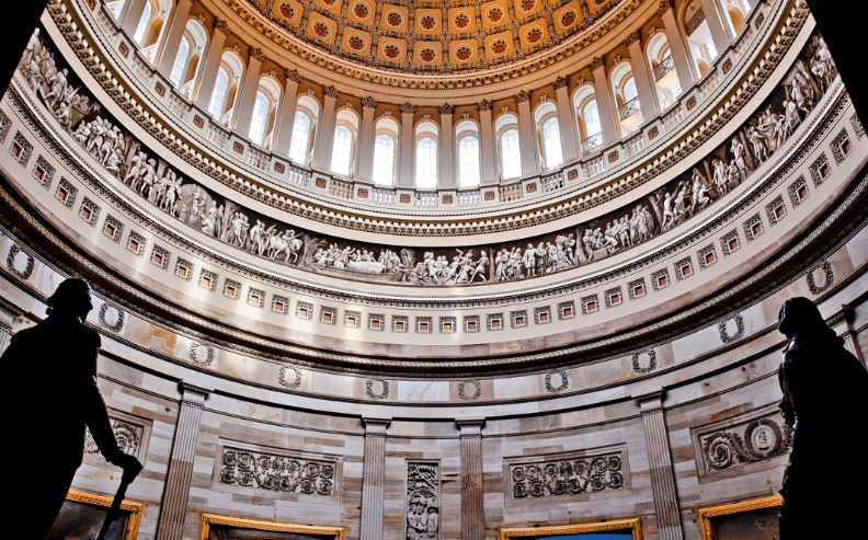 Interior view of a grand rotunda with ornate architectural details, statues silhouetted in the foreground, large arched windows, and intricate artwork decorating the upper walls and dome ceiling.