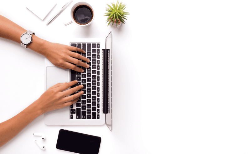 A person typing on a laptop at a white desk, surrounded by a notebook, pen, coffee cup, small plant, smartwatch, wireless earphones, and a smartphone. The right side of the desk is empty.
