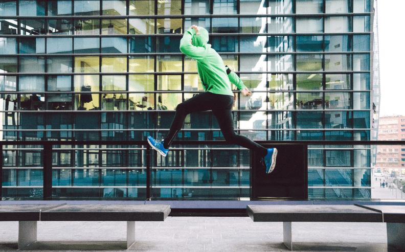 A person in a green hoodie, black pants, and blue sneakers is mid-air, leaping between two benches outdoors, with a modern glass building in the background.
