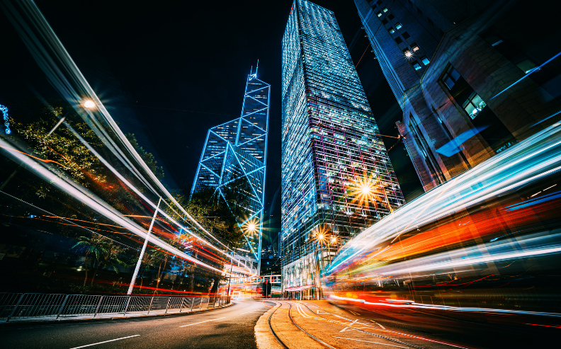 City street at night with tall, illuminated skyscrapers and light trails from passing vehicles, creating a vibrant, dynamic scene with reflections on the glass buildings and glowing streetlights.