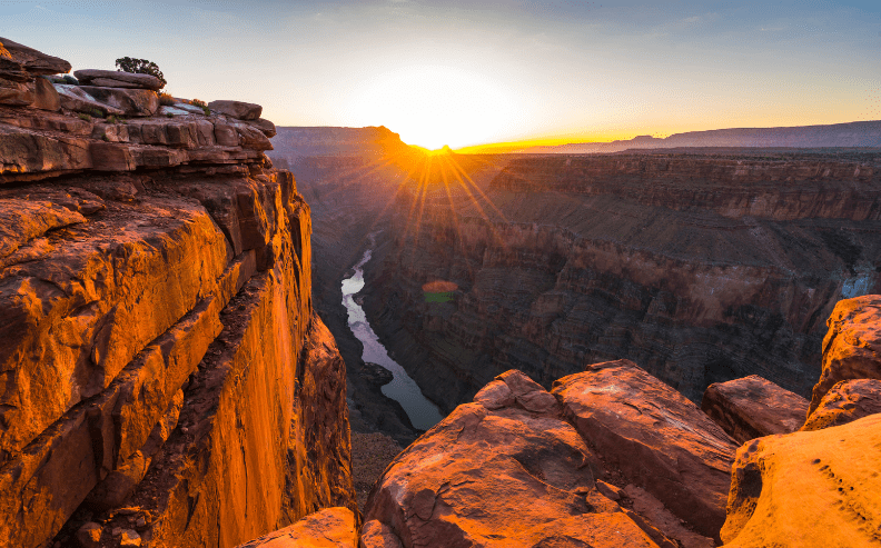 Sunrise over the Grand Canyon, with golden sunlight illuminating the rocky canyon cliffs and a winding river flowing through the deep gorge below.