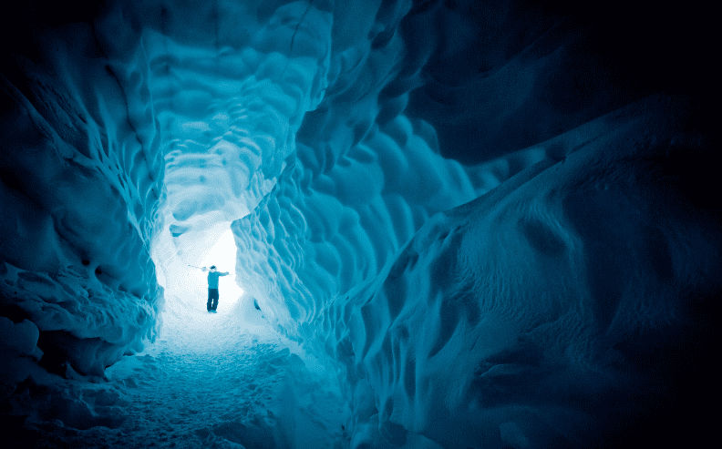 A person stands at the illuminated entrance of an icy cave with sculpted, blue walls, casting dramatic shadows. The textured ice creates a tunnel effect leading toward the bright opening.
