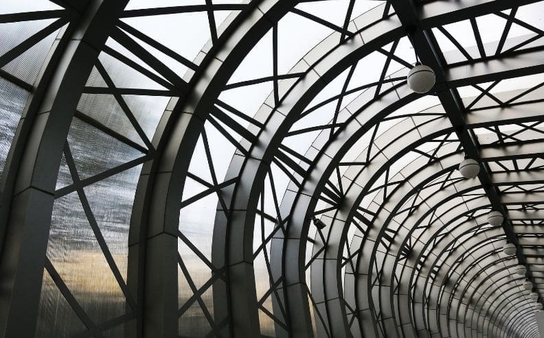 Curved steel and glass architectural structure with intersecting metal beams forming a tunnel-like roof, lit by several round ceiling lights, with natural light filtering through the panels.