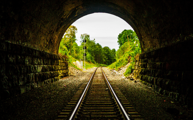 View of railroad tracks emerging from a dark tunnel into a green, tree-lined landscape under a cloudy sky, with signal lights visible in the distance.
