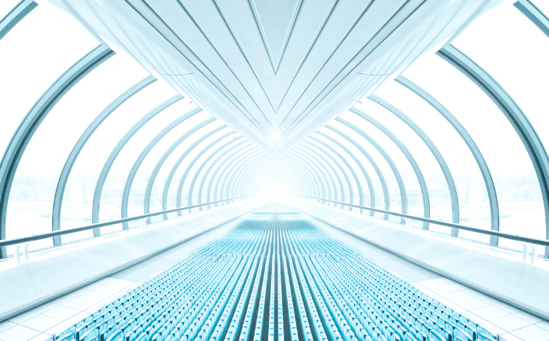 A futuristic, tunnel-like walkway with arched glass and metal beams, illuminated by bright white light at the far end, creating a sense of depth and symmetry. The floor features blue, patterned panels.